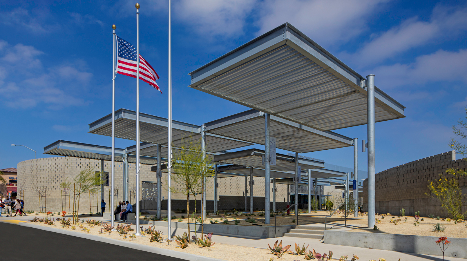 Photo of the CAB pedestrian plaza at Otay Mesa. It features desert landscaping, multiple staggered canopies, and a low tan building in the background. There are three large flagpoles in the foreground with an U.S. flag flying on one.