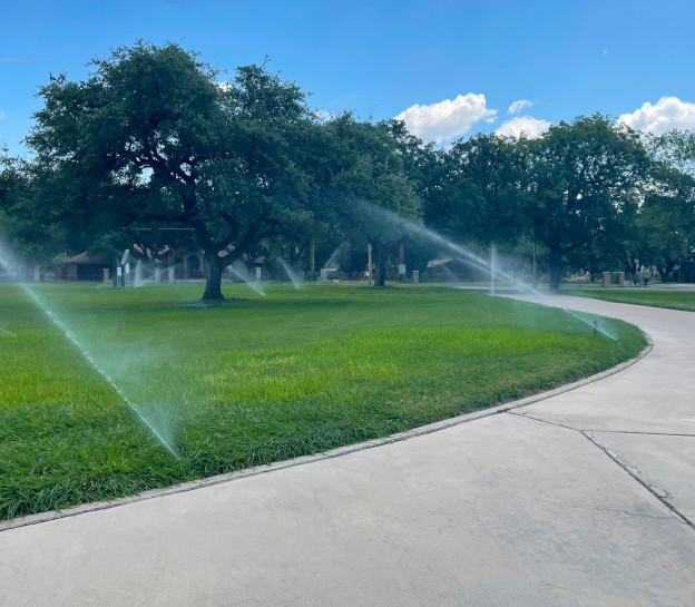 A view of large lawn with several large trees and multiple in-ground sprinklers watering the grass. A cement path curves around the lawn. Taken by Johnny Ferraro (7PMD).