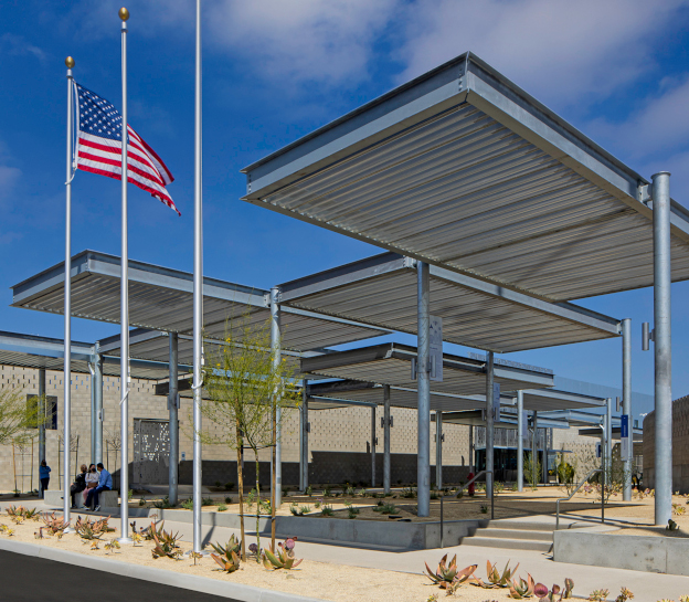 Photo of the CAB pedestrian plaza at Otay Mesa. It features desert landscaping, multiple staggered canopies, and a low tan building in the background. There are three large flagpoles in the foreground with an U.S. flag flying on one.