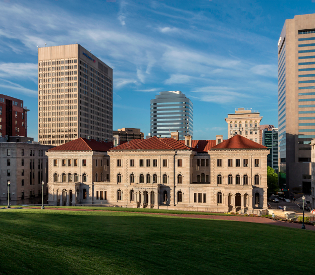 A photo of the Lewis F. Powell, Jr. Courthouse with several downtown Richmond buildings in the background and a grass lawn in the foreground. The building is three stories tall and in the Italianate style with white stone walls and a red tile roof.