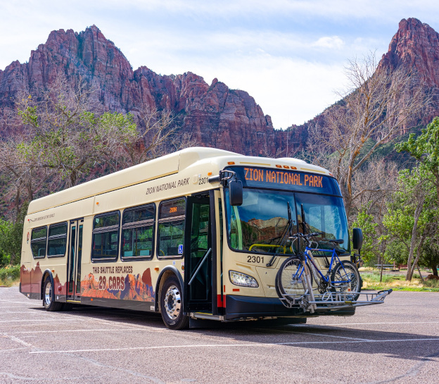 Picture of a full-size Zion National Park Bus in a parking lot with the mountains of the park in the background. The bus has a bike rack on the front and says Zion National Park on the front banner. On the side it says 