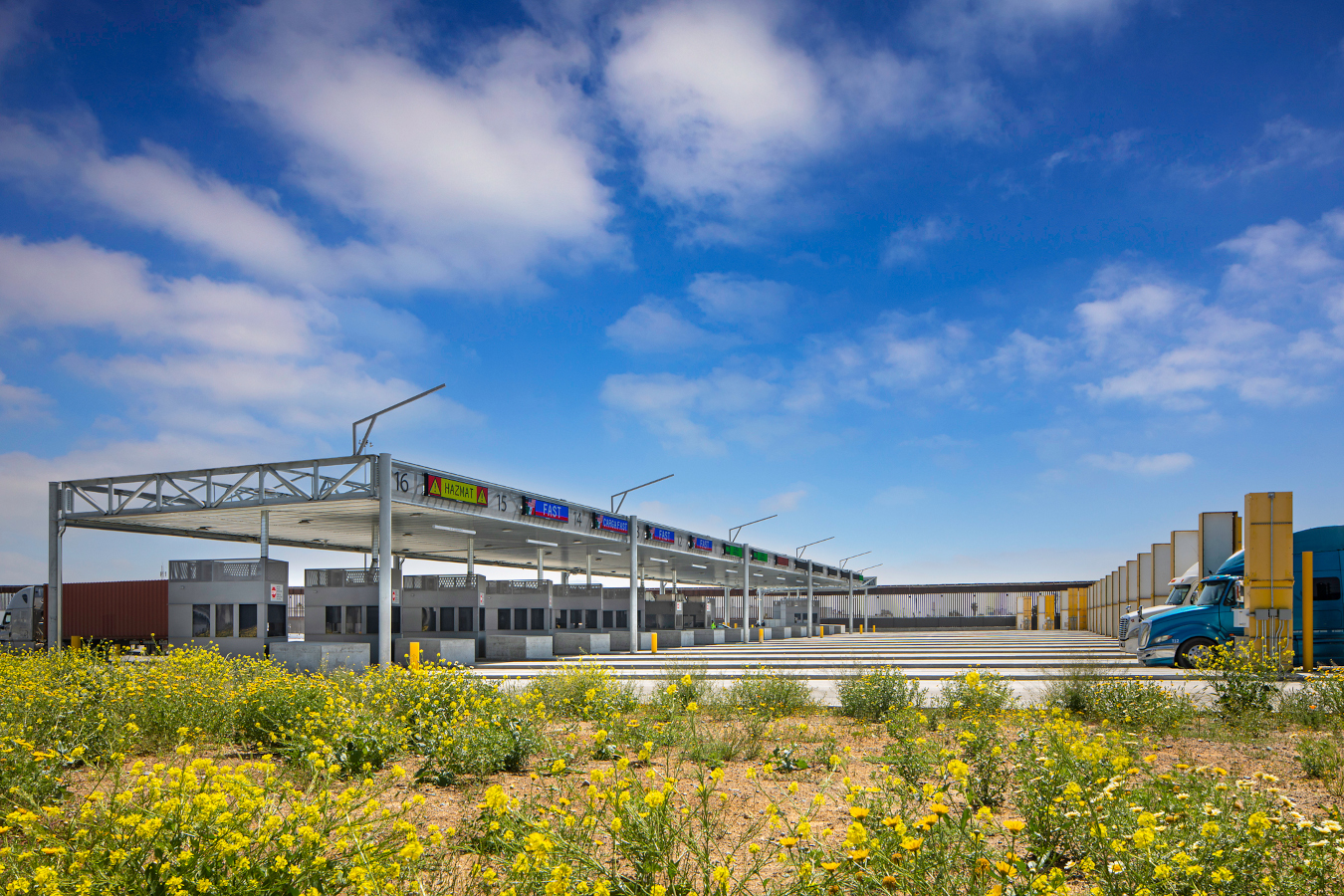 Photo of the import primary inspection booth at Otay Mesa. It features multiple truck lanes going horizontally across the image. On the right, there are two semi trucks in separate lanes. On the left is a single large canopy and inspection booths with number lanes that go up to 16. The closest lanes say Hazmat and Fast.